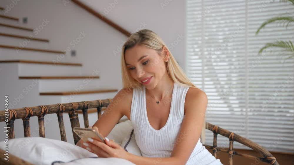 Charming blonde model in a white top chatting with her friends using a modern phone sitting indoor Charming blonde model in a white top chatting with her friends using a modern phone sitting indoor
