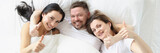 Two smiling women and man lie on bed and hold their thumbs up