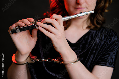 Woman musician with a flute in handcuffed hands on a studio dark background. Problems of musical playing at concerts, the concept of non-freedom and arrests