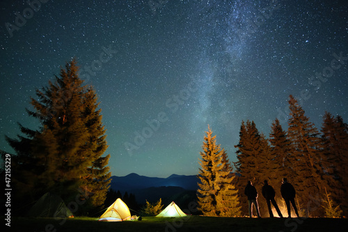 Hikers resting besides bright glowing bonfire near illuminated tourist tents on camping site in dark mountain woods under night sky with sparkling stars. Active lifestyle and outdoor living concept