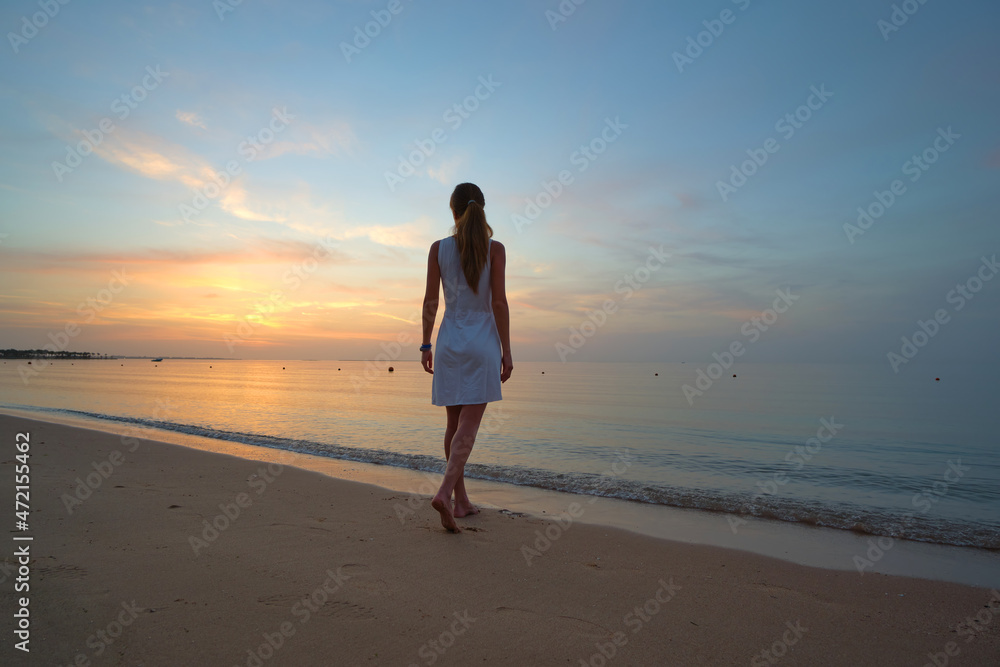 Lonely young woman standing on sandy beach by seaside enjoying warm tropical evening