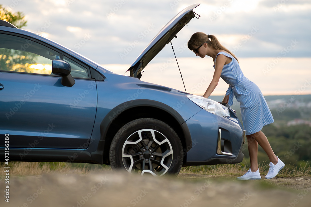 Stranded young woman driver standing near a broken car with popped up bonnet inspecting her vehicle motor
