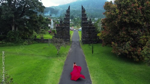 Pretty asian woman runs to balinese gates in long red dress in slowmotion on Hangara gates in Bali Indonesia. fashion concept. High quality fullhd aerial footage