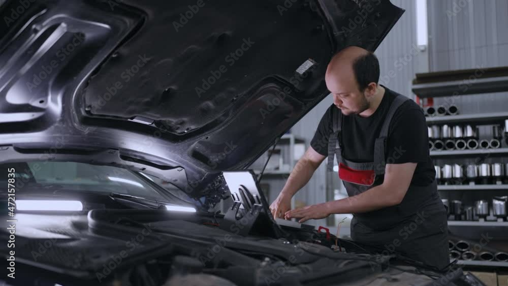 A male auto repairman in a car service with a computer checks the hood of the car A male auto repairman in a car service with a computer checks the hood of the car