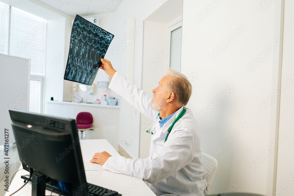 Side view of serious senior adult male doctor examining MRI spine image of patient sitting at desk with computer in medical office room, thinking about diagnosis, expressing concern of illness.