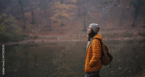 Young woman traveler in a yellow jacket and hat with a backpack breathing fresh air in the autumn forest
