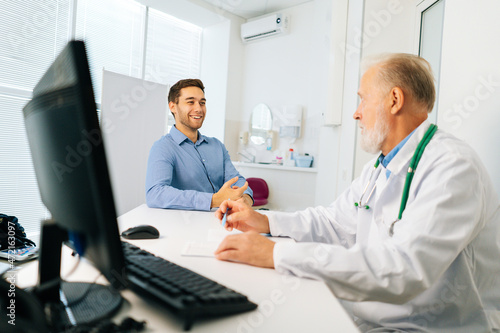Happy male patient talking with mature adult doctor sitting at table during checkup visit in medical clinic office. Handsome young man client listening to older senior physician explaining treatment.