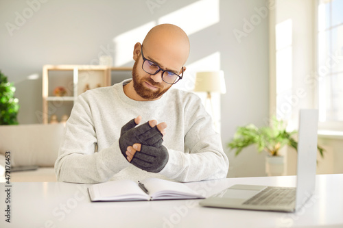 Unhappy man in glasses sitting at desk and looking at his aching hands in compression gloves. Man suffering from acute rheumatoid arthritis pain wears special fingerless gloves for working on computer photo