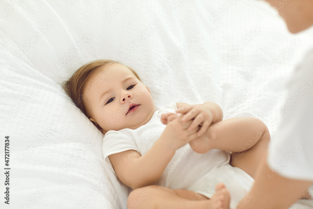 Cute baby boy or girl with mother. Beautiful little child resting on soft white cotton duvet in comfortable infant bed, playing with his or her bare feet and looking at mom while she's changing diaper Cute baby boy or girl with mother. Beautiful little child resting on soft white cotton duvet in comfortable infant bed, playing with his or her bare feet and looking at mom while she's changing diaper