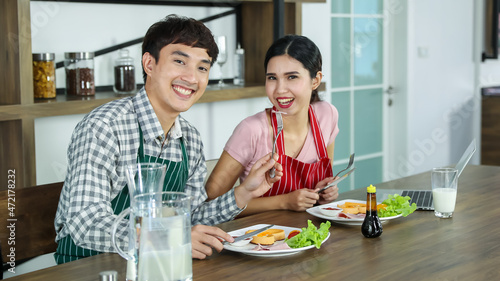 Romantic young Asian lovers happy talking  enjoy cooking healthy food together by spreading butter on toasted bread  chopping organic onion on cutting board on kitchen table of frying pan
