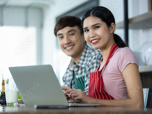 Beautiful Asian girl smiling as happy to study online food recipe on laptop together with chef boyfriend
