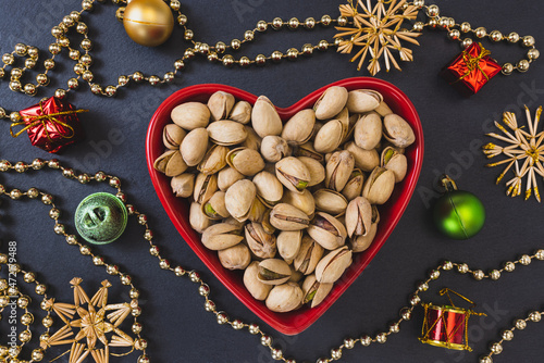 Roasted salted pistachios shelled in heart shaped red bowl on black background with Christmas decoration, concept of healthy eating vegan food and snack on winter holidays. Close up, selective focus