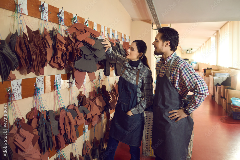 Shoe factory workers standing near wall of peg hangers with cut out natural leather details of future boots. Two young colleagues talking about footwear making process. Manufacturing industry concept