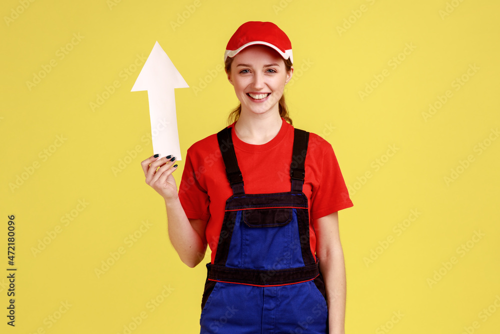 Portrait of attractive worker woman standing and looking at camera with happy expression, holding white arrow pointing up, career growth. Indoor studio shot isolated on yellow background.