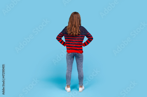 Full length back portrait of woman wearing striped casual style sweater, posing backwards with hands on hips, standing calm unrecognizable, waiting. Indoor studio shot isolated on blue background.