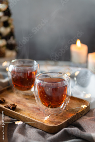 Winter hot tea in glass cup with steam in cozy composition. Two cups of black tea with steam on wooden tray on plaid with candles and Christmas tree. Close up. Christmas menu, greeting card