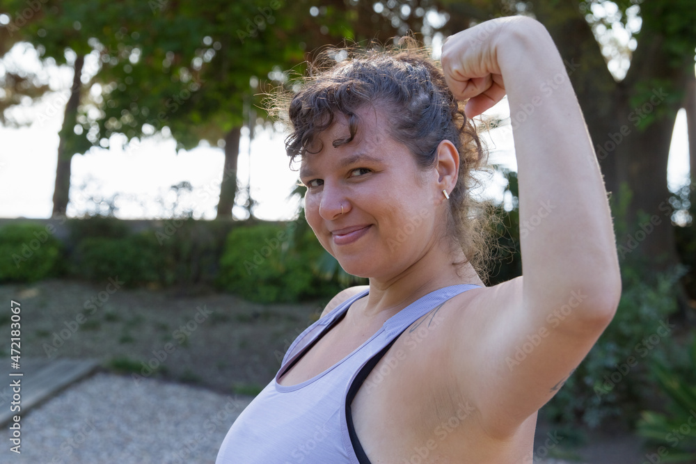 Portrait of cheery woman with curly hair doing sports. Chubby woman in blue top with tattoos proudly looking at camera, jokingly showing muscles. Sport, body positive concept Portrait of cheery woman with curly hair doing sports. Chubby woman in blue top with tattoos proudly looking at camera, jokingly showing muscles. Sport, body positive concept