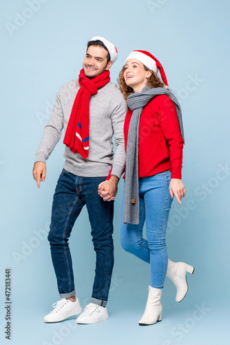 Full length portrait of Caucasian couple wearing Christmas outfits holding hands and looking up in light blue isolated studio background
