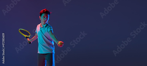 Cropped portrait ofd boy, tennis player training, serving ball isolated over blue background in neon lights. Flyer © Lustre