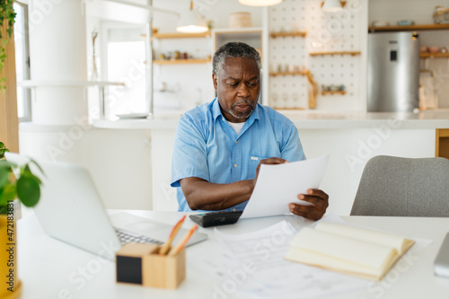 African senior man sitting at home and looking at bills he has to pay. He is paying it online over a laptop.
