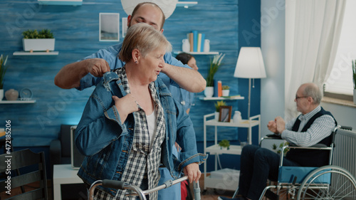 Slika na platnu Disabled woman getting dressed with jacket while nurse giving assistance in nursing home