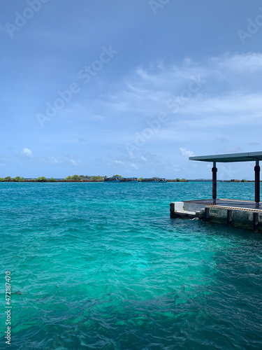A turquoise water jetty in the Maldives overlooking an island with docked ships. © Elena