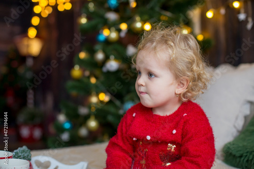 Cute toddler child, curly blond girl in a Christmas outfit, playing in a wooden cabin on Christmas, decoration around her. Child reading book and drinking tea
