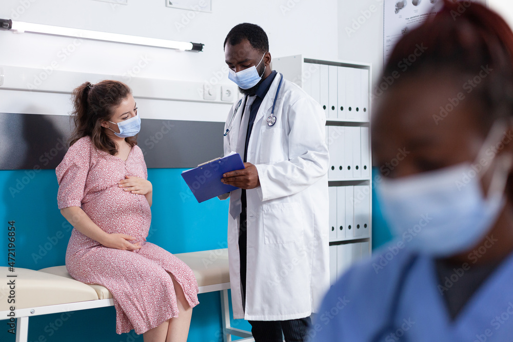Physician showing checkup papers to pregnant woman in medical office. General practitioner doing examination with patient expecting child to check healthcare while wearing face masks Physician showing checkup papers to pregnant woman in medical office. General practitioner doing examination with patient expecting child to check healthcare while wearing face masks