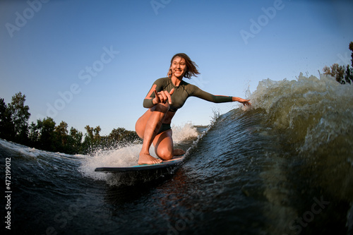 smiling woman sits on surf rides and touches waves with one hand and show hand gesture