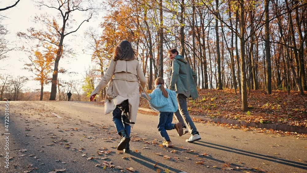 Young parents running by paved road in autumn park with little daughter jumping hanging on their hands. Slow motion, tracking shot. Back view Young parents running by paved road in autumn park with little daughter jumping hanging on their hands. Slow motion, tracking shot. Back view