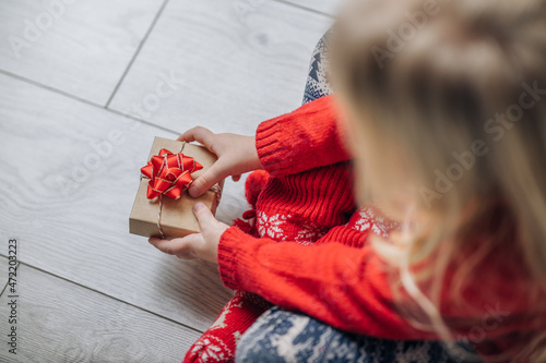 Child hands holding one gift box wrapped in kraft paper tied with red ribbon boow over wooden background. Top view, place for text. Holiday concept. photo