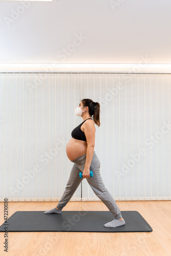 A pregnant woman with face mask standing doing dumbbell exercises at home