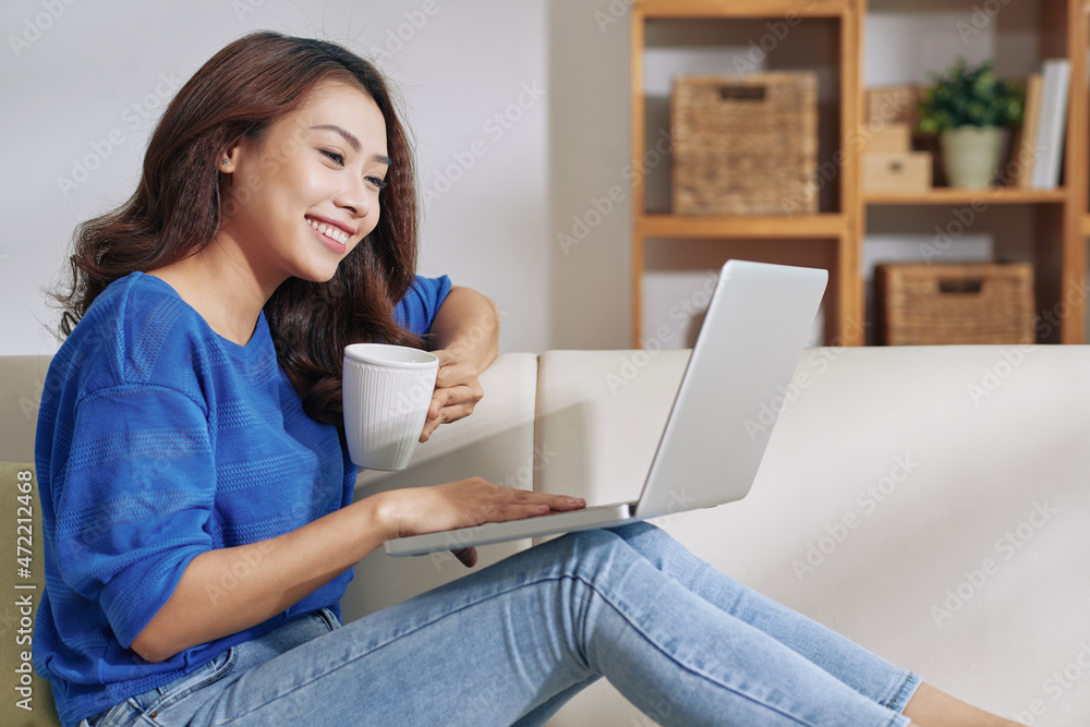 Smiling young Asian woman sitting with laptop on knees and drinking coffee while working at home Smiling young Asian woman sitting with laptop on knees and drinking coffee while working at home