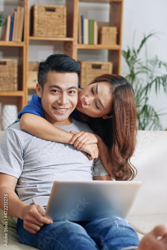 Beautiful Asian girl embracing boyfriend and kissing him while her using laptop at home © DragonImages