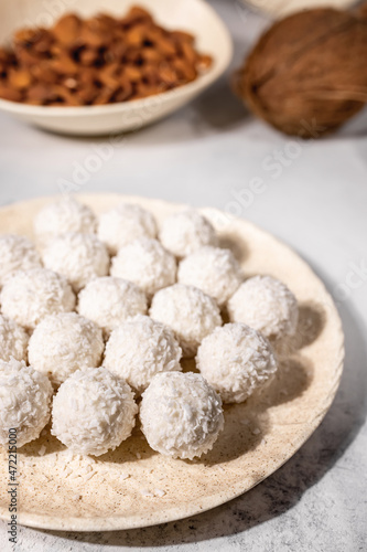White sweet coconut candies in plate, coconuts and almond nuts on background, selective focus, vertical