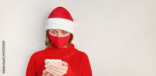 Christmas Woman in santa's hat, red sweater and wearing red medical nask stares surprised at phone's screen. Online shopping.Sale and discount concept.Mockup photo