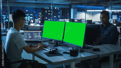 Night Office: Young Japanese Man in Working on Green Screen Chroma Key Desktop Computer. Team of Programmers Typing Code, Creating Modern Software, e-Commerce App Design, e-Business Programming