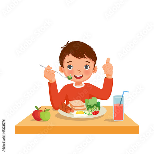 Cute little boy eating breakfast with bread, fried egg, broccoli, vegetables holding fork with sausage showing thumb up gesture.

