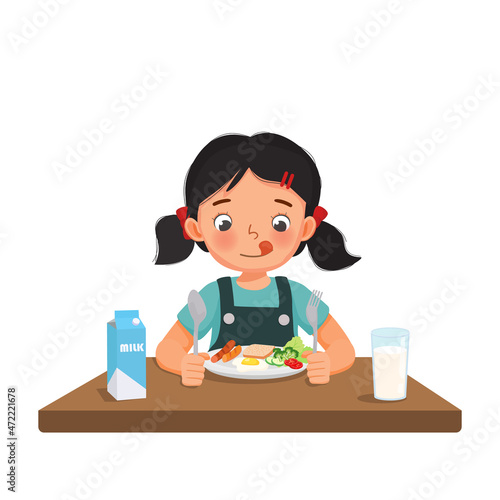 Cute little girl feeling excited eating breakfast with bread, fried egg, broccoli, vegetables, milk holding spoon and fork.