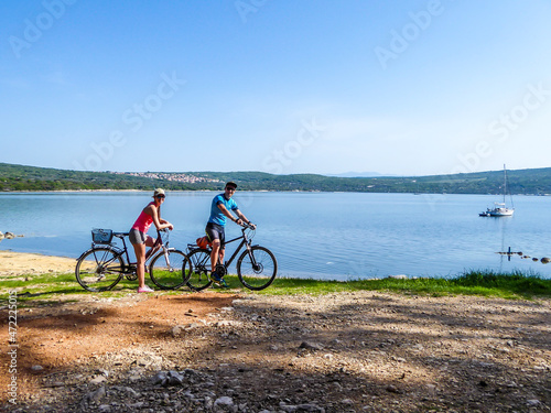 A couple in sporty outfits stand on their bikes by the seashore and admire the view behind them. Coast is partially sandy, stony and grassy. The water in the bay is calm, boat anchored to the shore