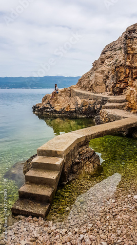 A girl sitting on a cliff emerging from the water. There is a pathway leading to the viewing point, over the sea. Water in the bay is calm and crystal clear. Stony beach. Big overcast.