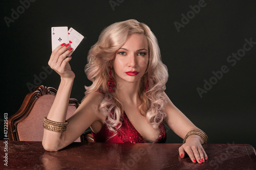 A gorgeous blonde woman in a bright red evening dress sits at a table with cards, holding in her hand aces cards and looking at the camera with an attentive gaze. Blurred details