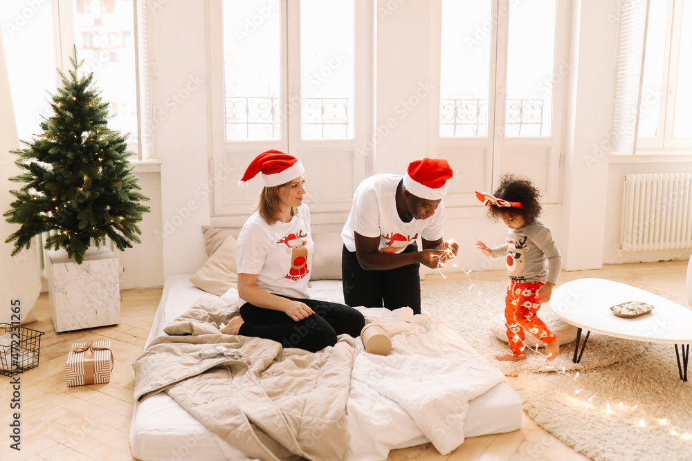 Fototapeta premium A multiracial mixed race family with a child a little girl in pajamas and Santa Claus hats are having fun preparing for the holiday decorating a Christmas tree in a cozy indoor house. Selective focus