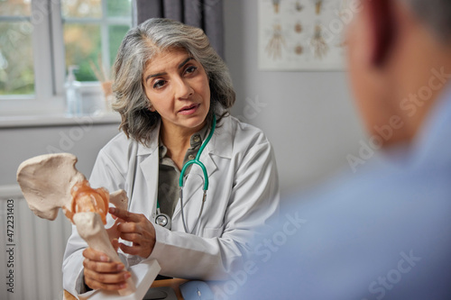 Mature Female Doctor Meeting With Male Patient Discussing Joint Pain In Hip Using Anatomical Model