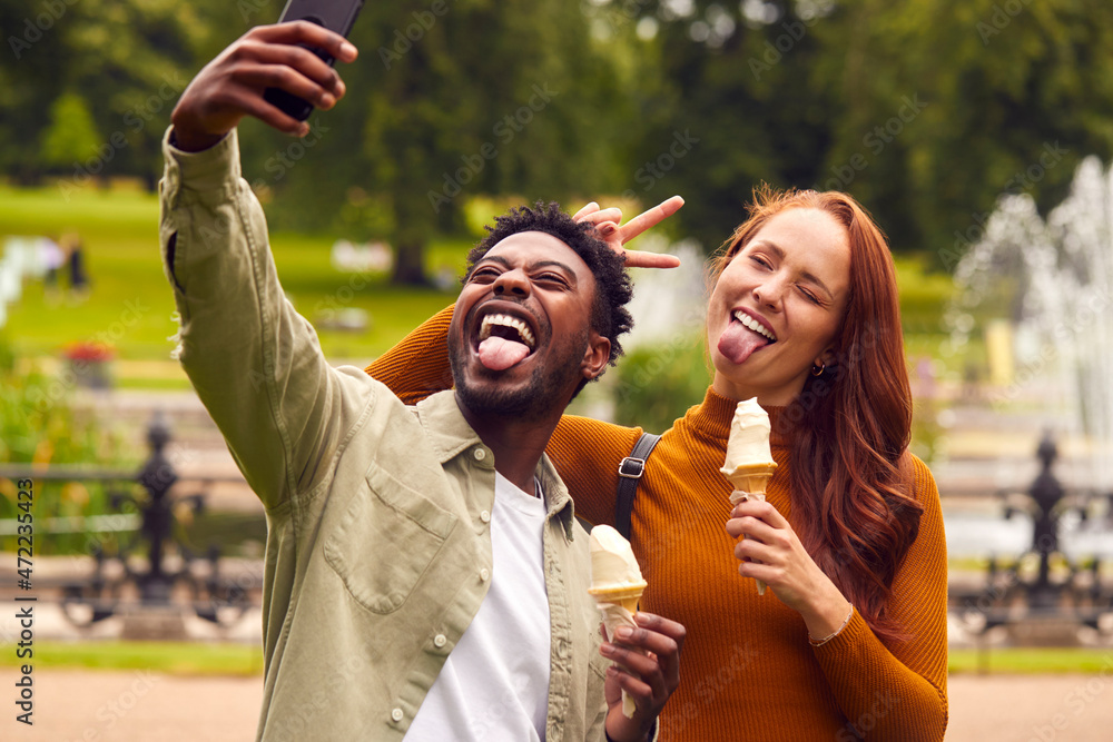 Obraz premium Young Couple Travelling Through City Together Eating Ice Creams In Park And Taking Selfie