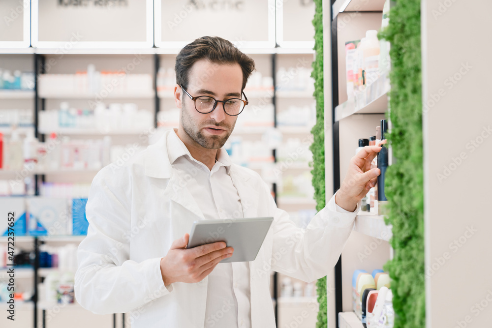 Male young handsome pharmacist druggist in medical white coat looking for drugs at prescription on digital tablet stretching to pills, vitamins, painkillers, medicines at pharmacy shelves in drugstore
