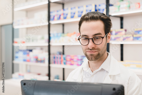 Caucasian male young pharmacist druggist working on computer, looking at screen in white medical coat looking for drugs, medicines, pills for customers in drugstore pharmacy