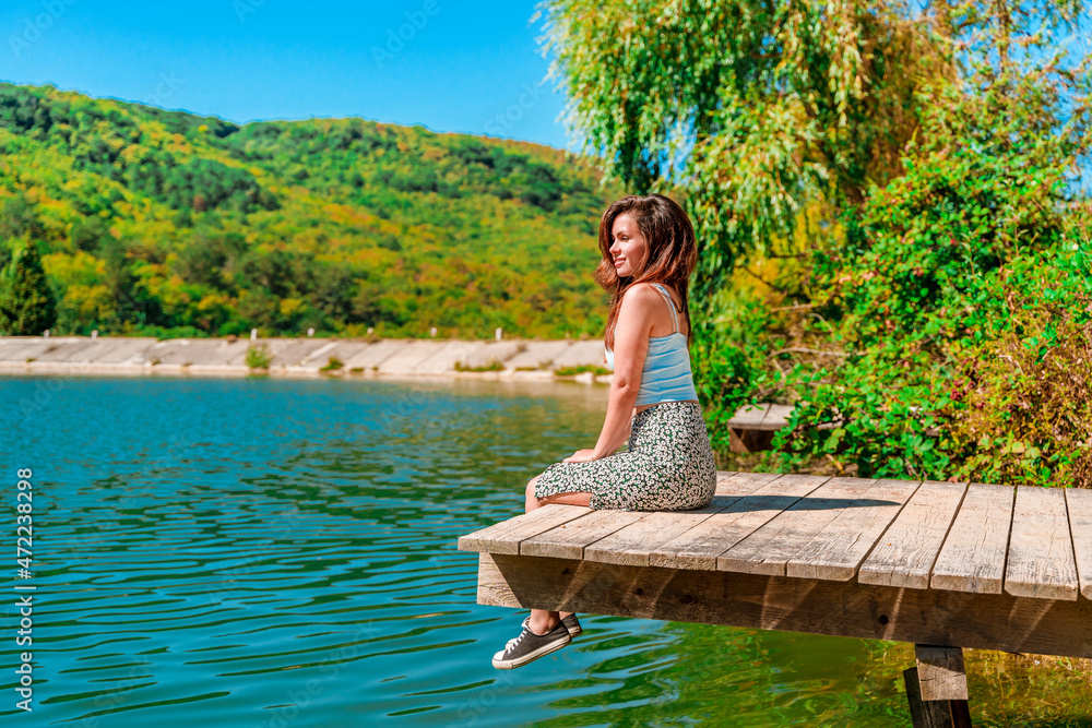 Fototapeta premium A charming young woman in a skirt is sitting on a wooden bridge against the background of nature with a lake in the forest. Travel, freedom, the concept of an active lifestyle.