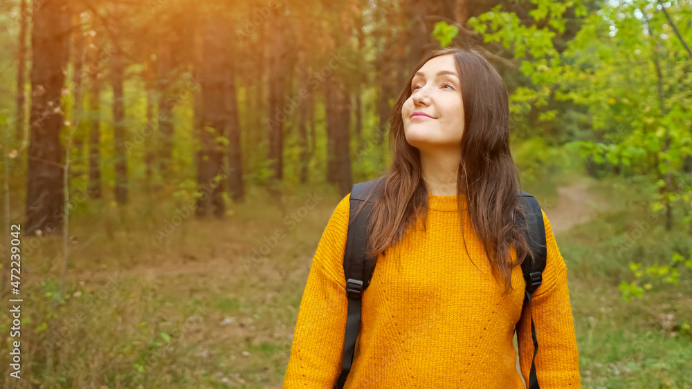 Happy dark haired lady hiker with backpack wearing warm orange sweater walks along empty narrow road running across green forest, sunlight Happy dark haired lady hiker with backpack wearing warm orange sweater walks along empty narrow road running across green forest, sunlight