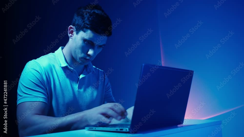 Man Works at a Laptop, Typing Text on the Keyboard. Indoors Neon Lighting and a Futuristic Atmosphere. Freelance and Remote Work Concept. Modern Technologies. Man Works at a Laptop, Typing Text on the Keyboard. Indoors Neon Lighting and a Futuristic Atmosphere. Freelance and Remote Work Concept. Modern Technologies.